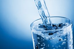 Das Bild zeigt ein Glas Trinkwasser, dass vor einem blauen Hintergrund mit einem Wasserstrahl befüllt wird.